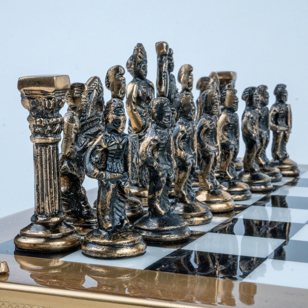 A set of ornate metallic chess pieces arranged on a checkered black-and-white board, photographed against a pale blue studio background.