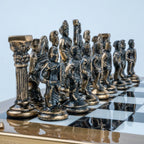 A set of ornate metallic chess pieces arranged on a checkered black-and-white board, photographed against a pale blue studio background.