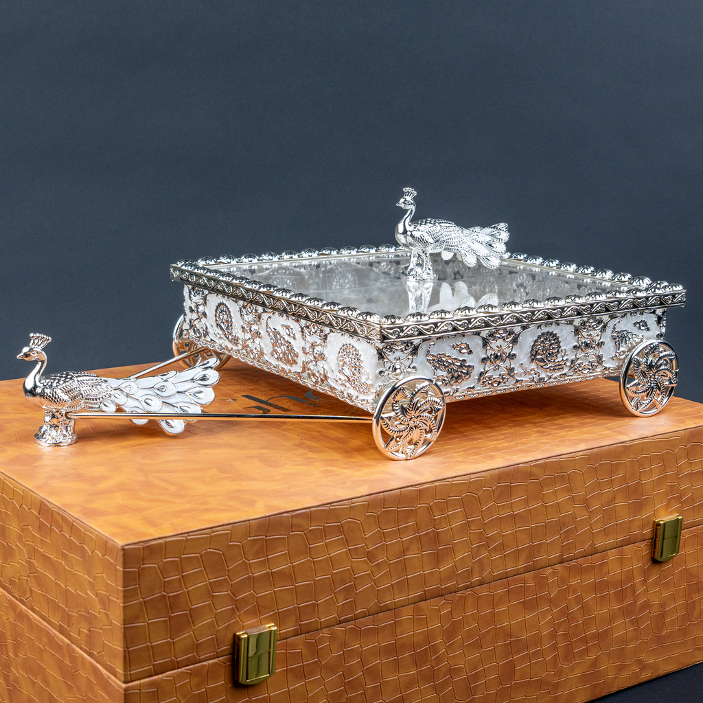 Silver decorative miniature horse-drawn chariot showpiece featuring an intricately detailed rectangular carriage with ornate embossed floral patterns, a transparent reflective top, decorative silver horse figurines, and four beautifully designed ornamental wheels. An elegant luxury home décor accent for tabletops, living rooms, display shelves and premium interiors.