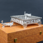 Silver decorative miniature horse-drawn chariot showpiece featuring an intricately detailed rectangular carriage with ornate embossed floral patterns, a transparent reflective top, decorative silver horse figurines, and four beautifully designed ornamental wheels. An elegant luxury home décor accent for tabletops, living rooms, display shelves and premium interiors.