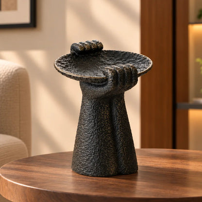 Dark textured sculptural pedestal bowl displayed on a round wooden table in a modern living room with warm natural lighting and a beige sofa.