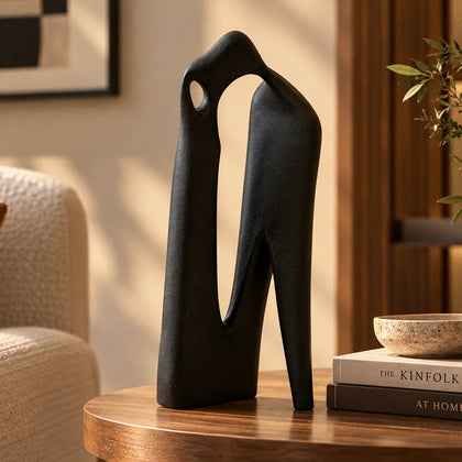 Tall matte-black abstract sculpture displayed on a round wooden side table in a modern living room, with stacked books, a textured bowl, greenery, and warm natural lighting.
