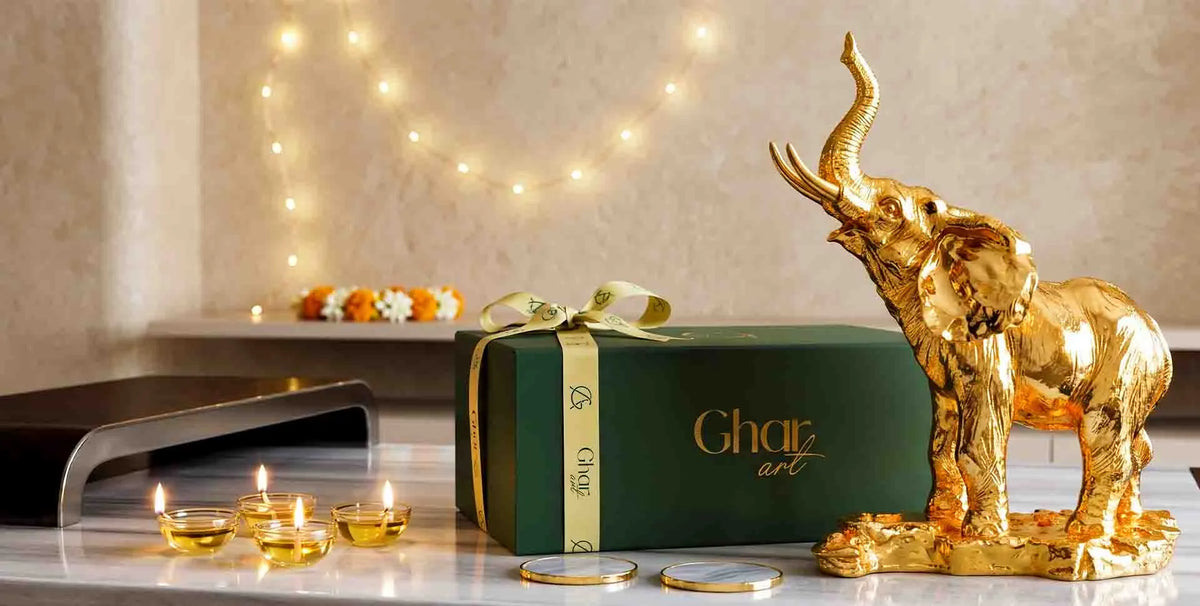 A dark green Ghat art gift box with a gold ribbon and bow, displayed next to a shiny gold elephant figurine on a marble surface, with lit candles and warm string lights in the background.