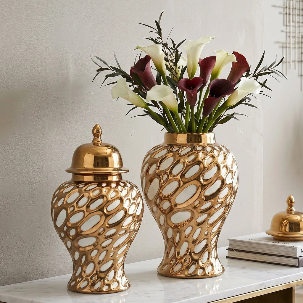 Two decorative gold ceramic vases with an open, bubble-like pattern sit on a marble-top console table. One vase is closed with a matching gold lid, and the other holds white and burgundy calla lilies.