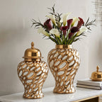Two decorative gold ceramic vases with an open, bubble-like pattern sit on a marble-top console table. One vase is closed with a matching gold lid, and the other holds white and burgundy calla lilies.