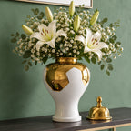 Ornate white and gold decorative vase filled with white lilies and green buds, displayed on a wooden surface against a deep green wall, with its lid placed beside it.