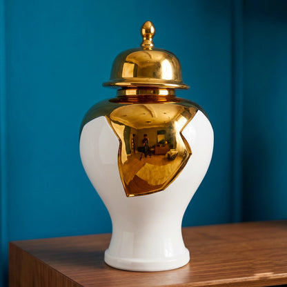 Ornate white and gold decorative vase displayed on a wooden surface against a blue wall, with its lid placed beside it.