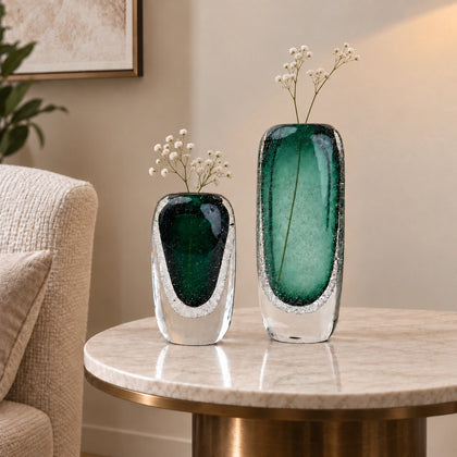 Two decorative glass vases in a styled home setting, one small and one large, both featuring a dark green and clear glass design with white baby's breath flowers inside, displayed on a marble-topped side table.
