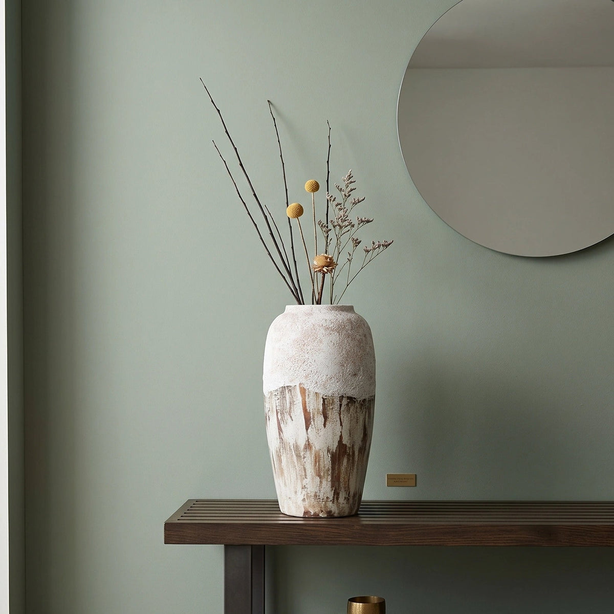 A tall textured ceramic vase with a white and brown mottled finish sits on a dark wooden console table in a styled room. The vase contains dried branches with small yellow accents.