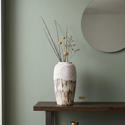 A tall textured ceramic vase with a white and brown mottled finish sits on a dark wooden console table in a styled room. The vase contains dried branches with small yellow accents.