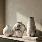 Three textured ceramic vases in off-white and gray with dark charcoal splatters, displayed on a light stone sideboard next to a window.