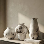 Three textured ceramic vases in off-white and gray with dark charcoal splatters, displayed on a light stone sideboard next to a window.