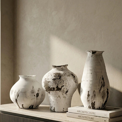 Three textured ceramic vases in off-white and gray with dark charcoal splatters, displayed on a light stone sideboard next to a window.