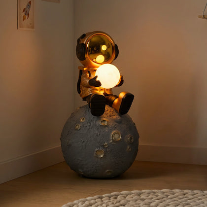 Gold astronaut figurine sitting on a large cratered moon, holding a glowing orb lamp with a chrome-gold visor.