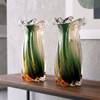 Two tall fluted glass vases with a green-to-clear gradient and amber accents, displayed on a wooden table against a light gray wall.