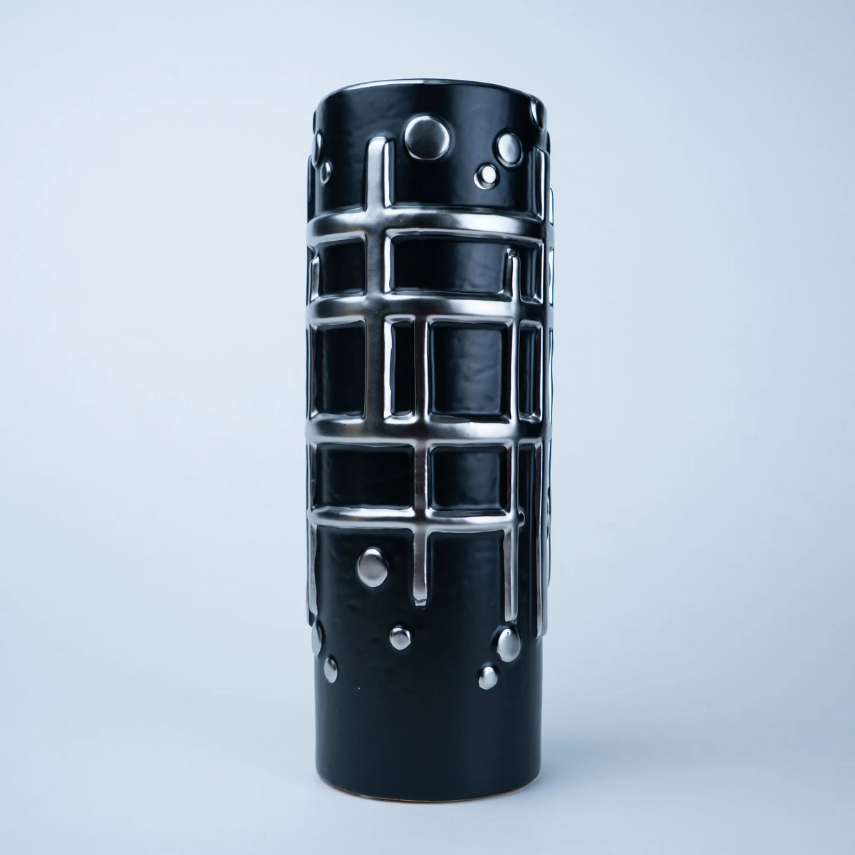 A black cylindrical decorative object with a glossy finish, featuring a raised silver grid pattern and silver circular studs, photographed against a light gray studio background.