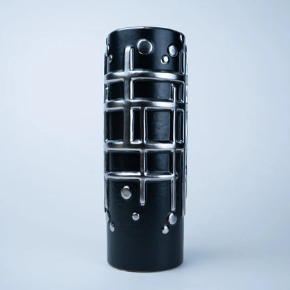 A black cylindrical decorative object with a glossy finish, featuring a raised silver grid pattern and silver circular studs, photographed against a light gray studio background.