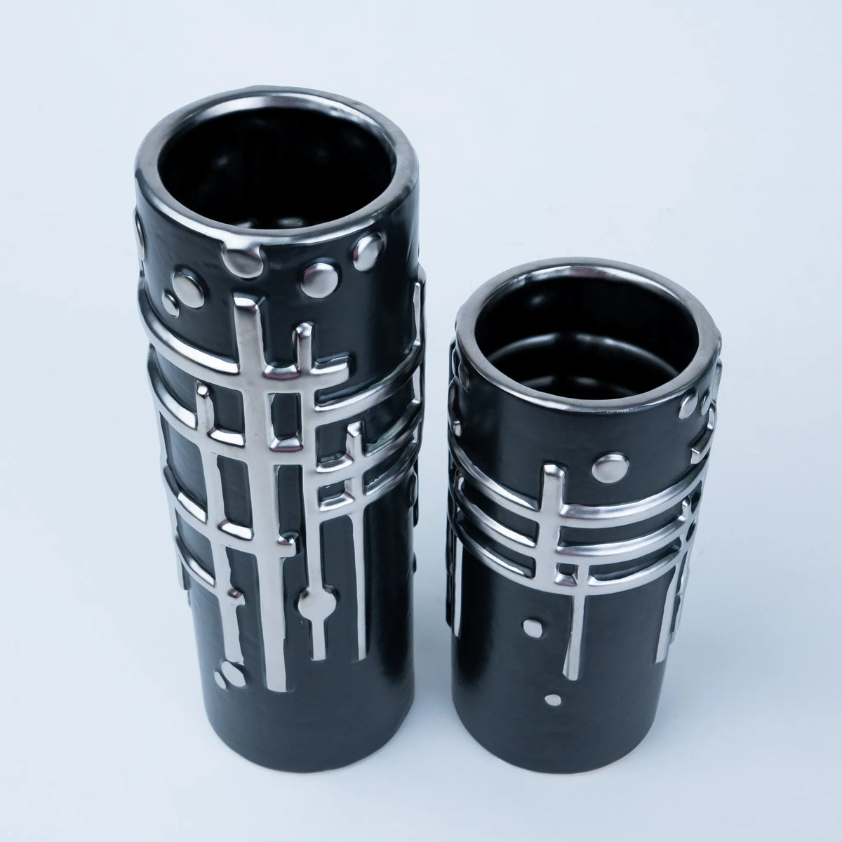 Two black cylindrical metal holders with silver geometric grid patterns, photographed on a light gray background.