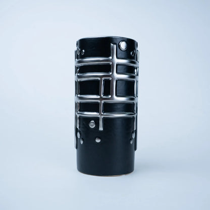 A black ceramic vase with a textured, ribbed body and raised silver metallic grid patterns, photographed against a clean, light gray studio background.