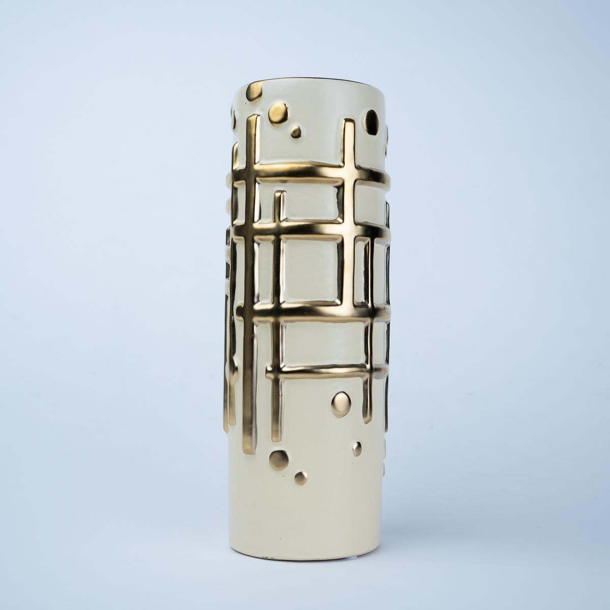 A modern cylindrical ceramic vase in cream with a gold geometric grid pattern and perforated details, set against a light blue-gray gradient background.