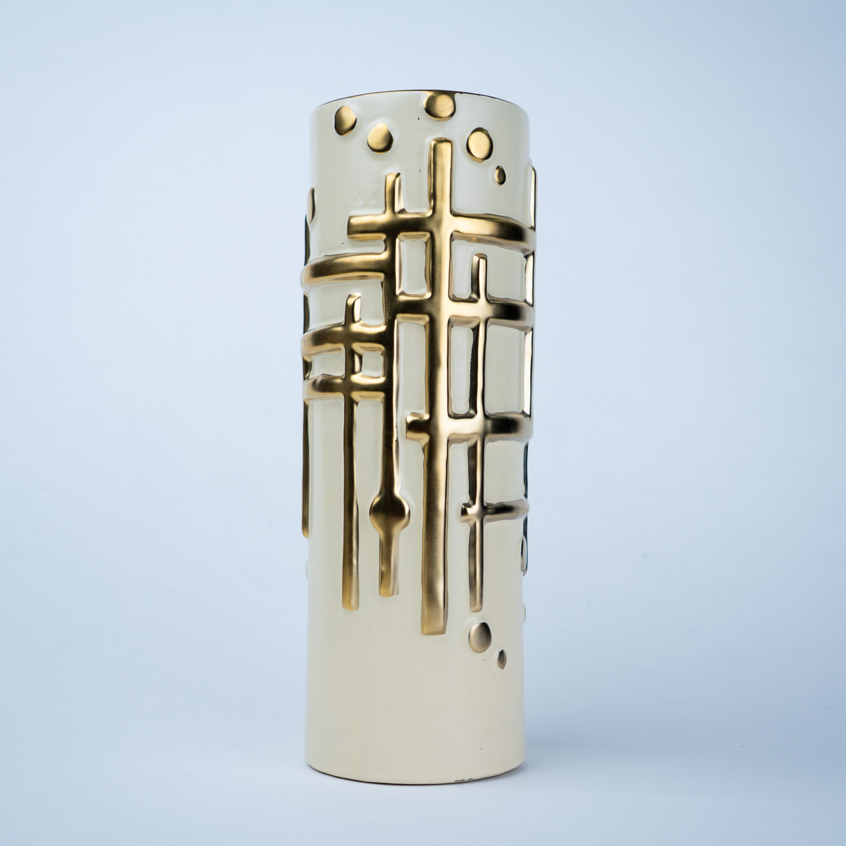 A cylindrical ceramic vase in cream with a gold geometric lattice pattern, photographed against a light blue-gray studio background.