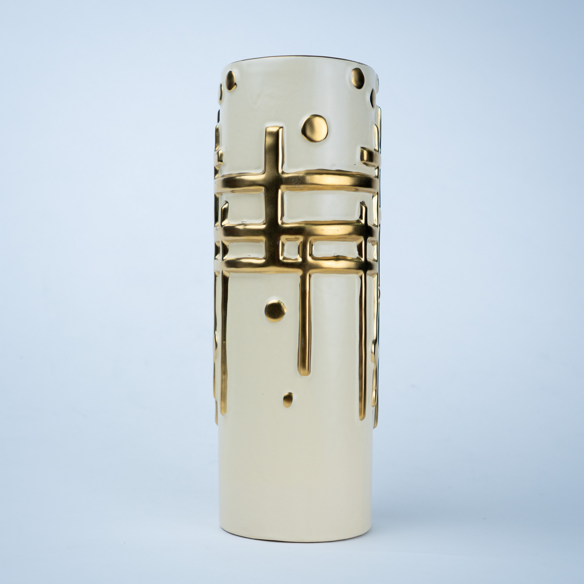 A cylindrical ceramic vase in a warm cream color with metallic gold geometric patterns, including a central cross motif and circular cutouts, set against a light blue gradient background.