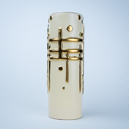 A cylindrical ceramic vase in a warm cream color with metallic gold geometric patterns, including a central cross motif and circular cutouts, set against a light blue gradient background.