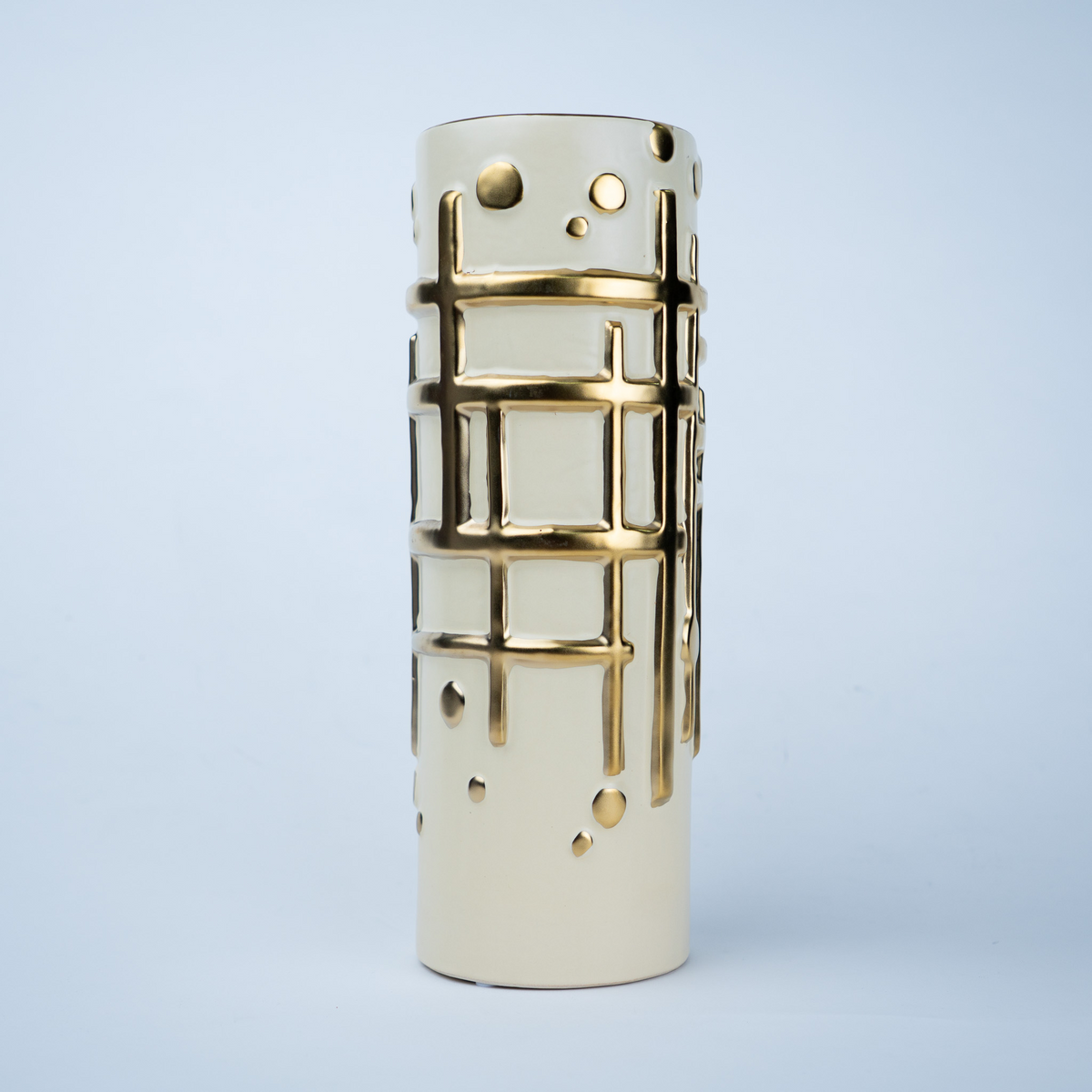 A modern cylindrical ceramic vase in cream with metallic gold cutout patterns of a grid and dots, set against a light blue background.