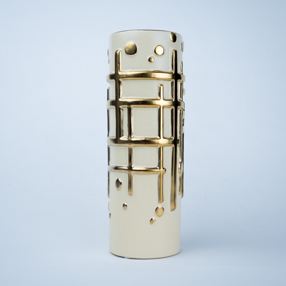 A modern cylindrical ceramic vase in cream with metallic gold cutout patterns of a grid and dots, set against a light blue background.