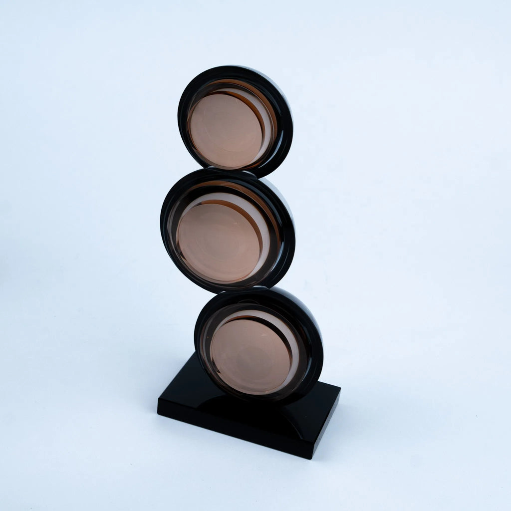 A modern abstract sculpture featuring three stacked circular glass shades in a warm taupe-brown color, mounted on a glossy black rectangular base against a clean white studio background.