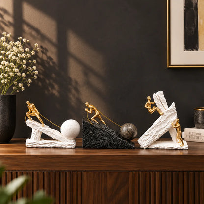 Three decorative sculptural figurines arranged on a wooden console table against a dark wall. Each figure is posed on a tilted stone-like base with a small ball, and the set includes white, black, and textured white versions.