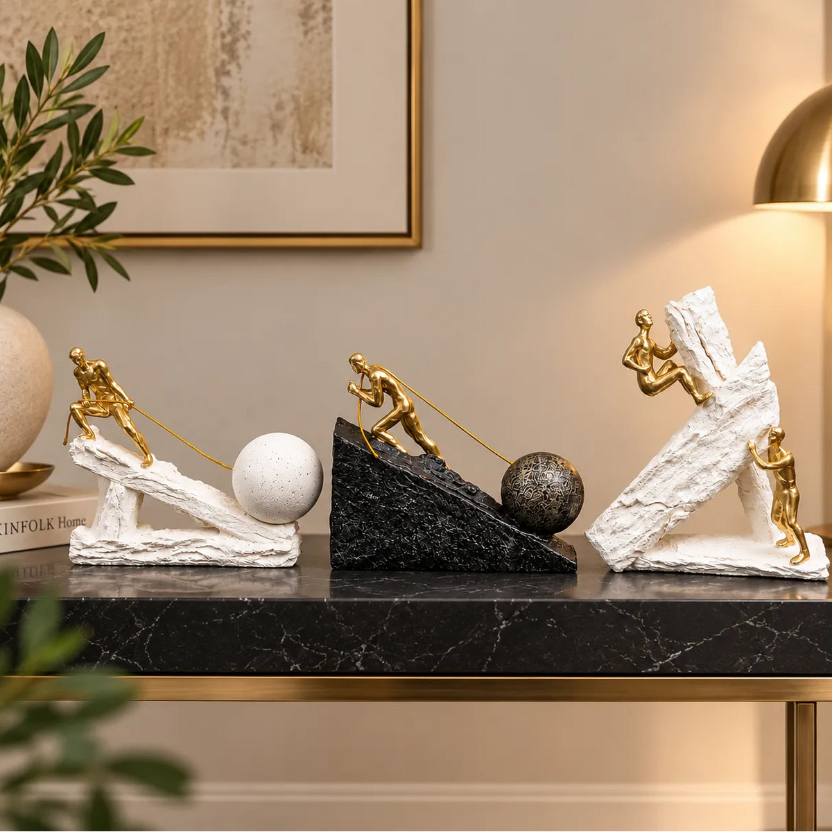 Three decorative sculpture sets on a black marble console table in a styled interior. The sets feature small gold human figures pushing white and black textured blocks with spheres, displayed against a beige wall with a lamp, framed art, and plants.