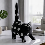 A black cat-shaped decorative object with white polka dots stands on a white tabletop in a softly lit room. The design includes white circular spots across the body and tail, with small white facial details.