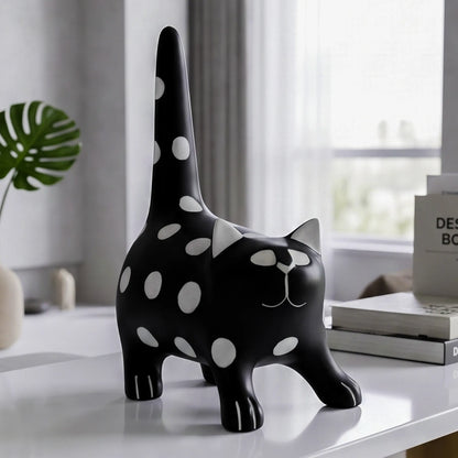 A black cat-shaped decorative object with white polka dots stands on a white tabletop in a softly lit room. The design includes white circular spots across the body and tail, with small white facial details.