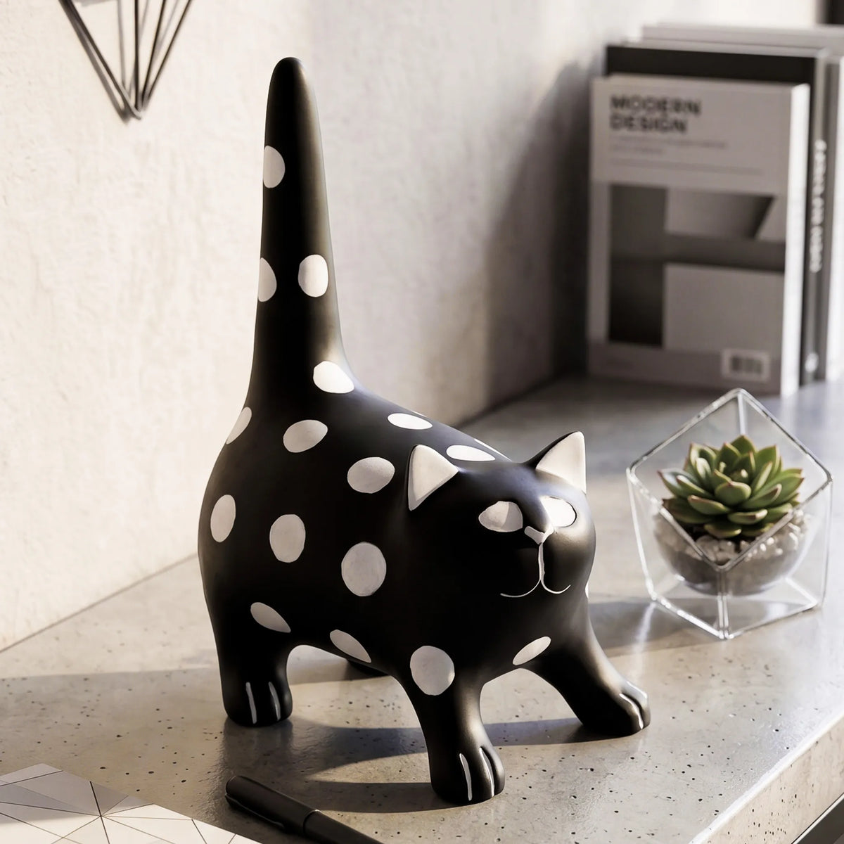 A black cat-shaped desk lamp or figurine with white polka dots, standing on a gray tabletop next to a small succulent in a glass terrarium.
