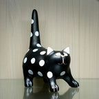 A glossy black cat figurine with white polka dots, white inner ears, and a white stitched-style face, standing on a reflective surface against a light wood background.
