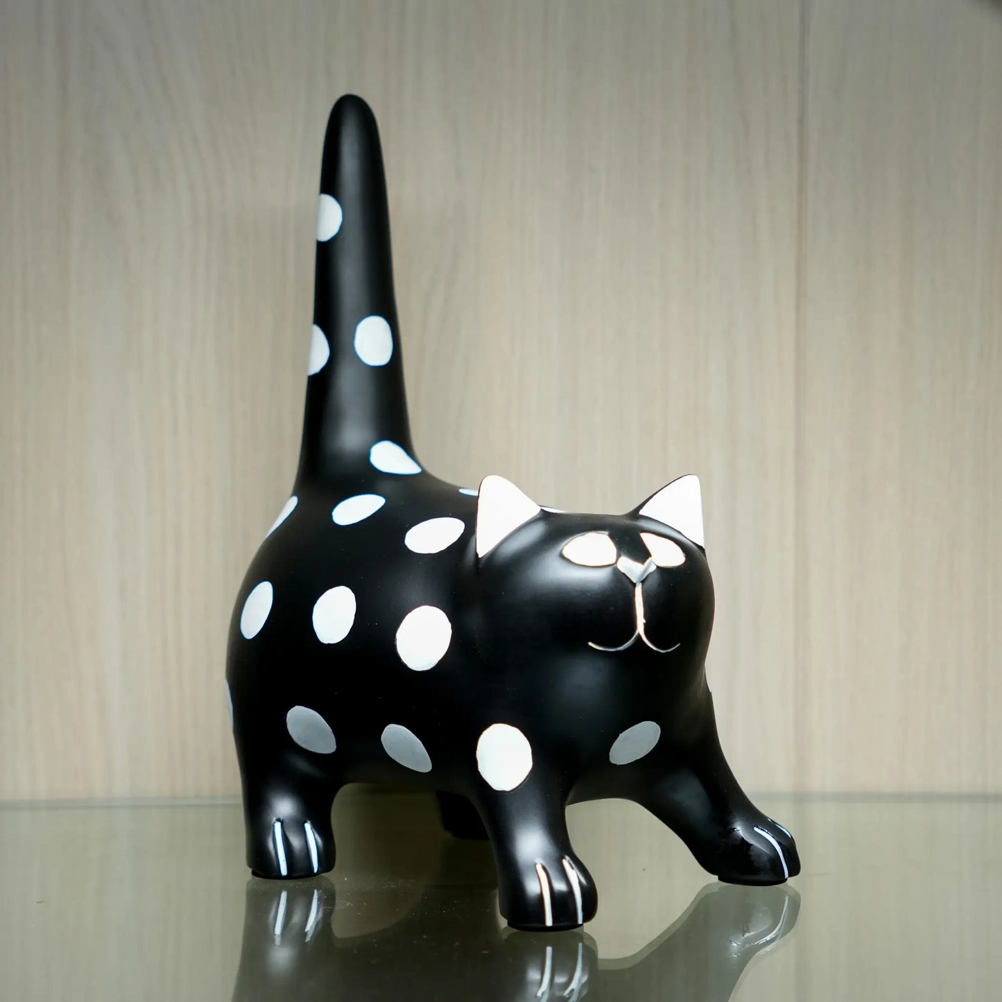 A glossy black cat figurine with white polka dots, white inner ears, and a white stitched-style face, standing on a reflective surface against a light wood background.