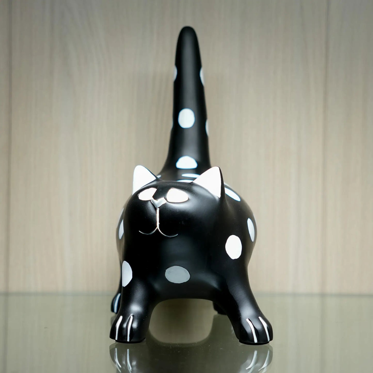A glossy black cat figurine with white polka dots, white inner ears, and white paw details, standing on a reflective surface against a light wood-paneled background.