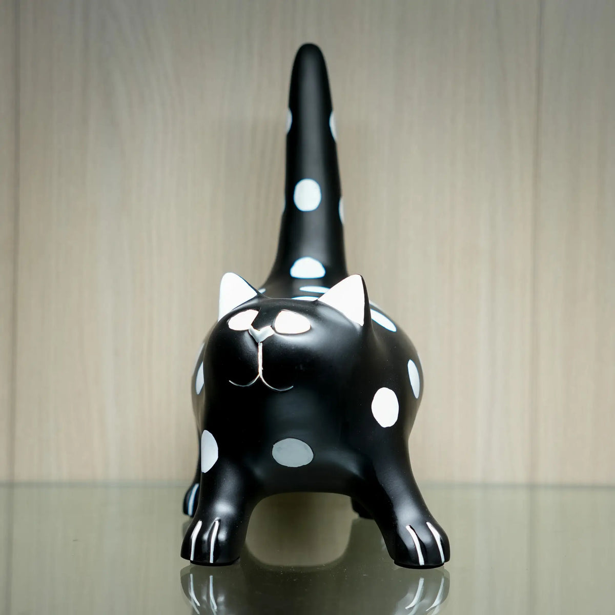 A glossy black cat figurine with white polka dots, white inner ears, and white paw details, standing on a reflective surface against a light wood-paneled background.