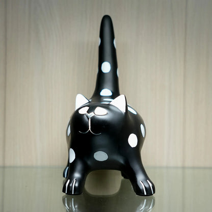 A glossy black cat figurine with white polka dots, white inner ears, and white paw details, standing on a reflective surface against a light wood-paneled background.