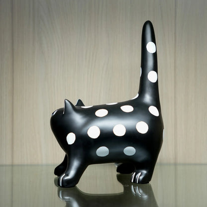 A glossy black cat figurine with large white polka dots, standing on a reflective surface against a light wood-paneled background.
