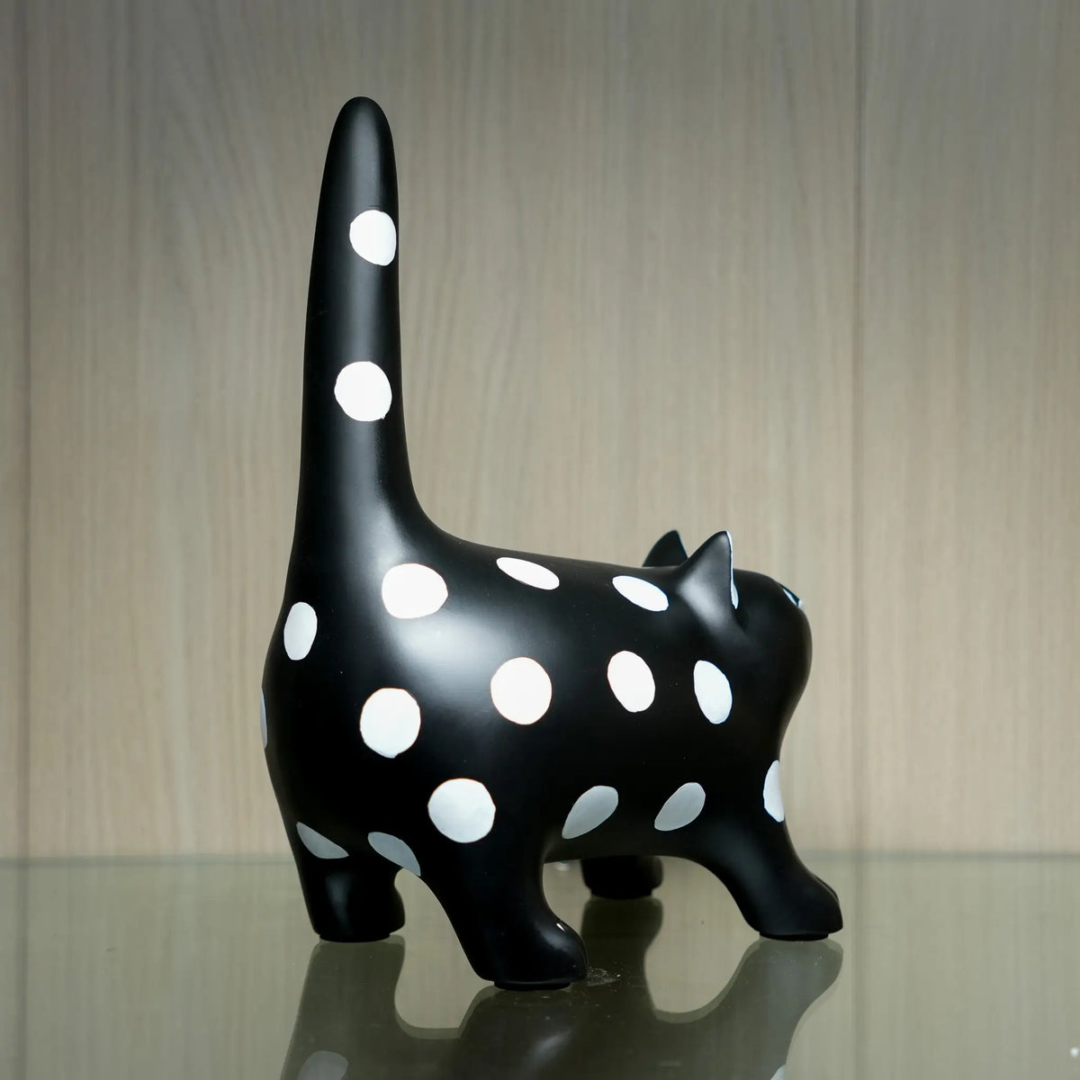 A glossy black cat figurine with large white polka dots, standing on a reflective surface against a light wood-grained background.
