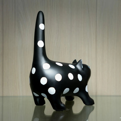 A glossy black cat figurine with large white polka dots, standing on a reflective surface against a light wood-grained background.