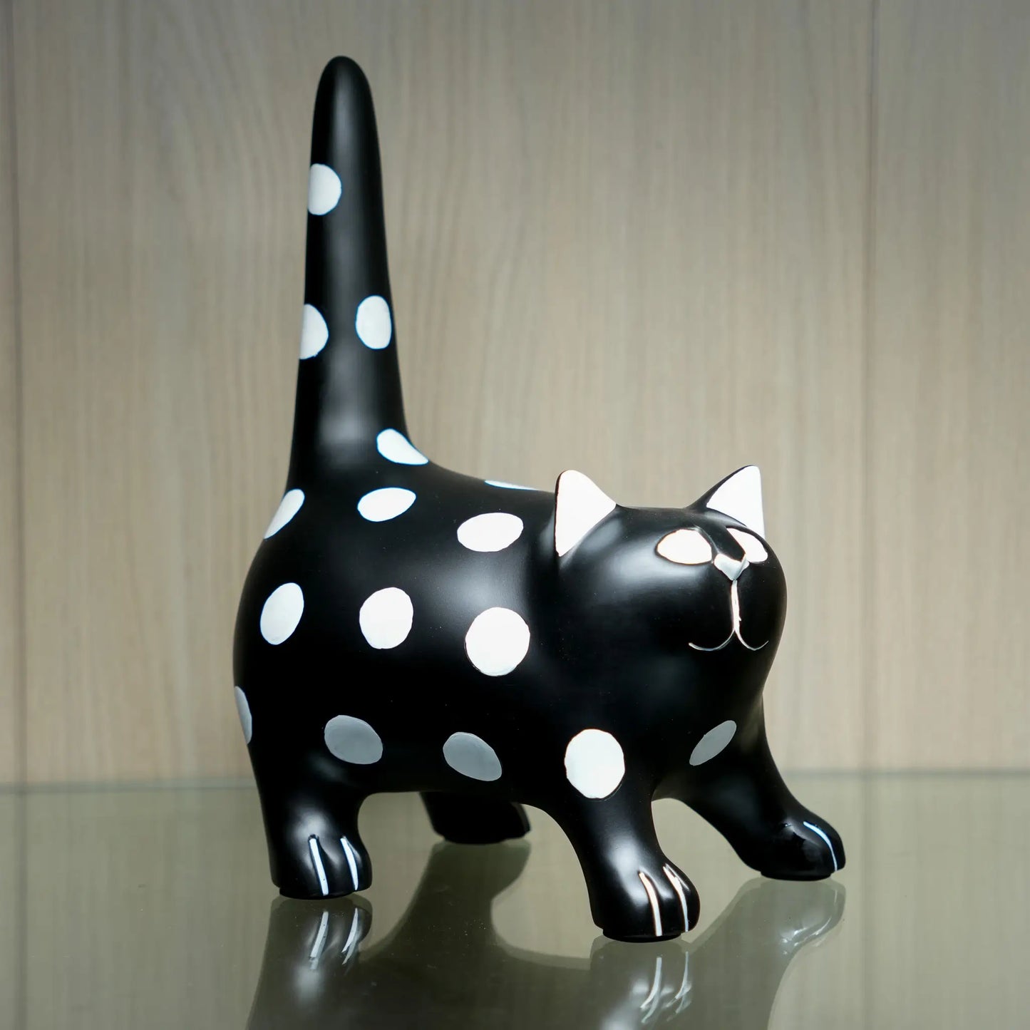 A glossy black cat figurine with white polka dots, white pointed ears, and white facial details, standing on a reflective surface against a light wood background.