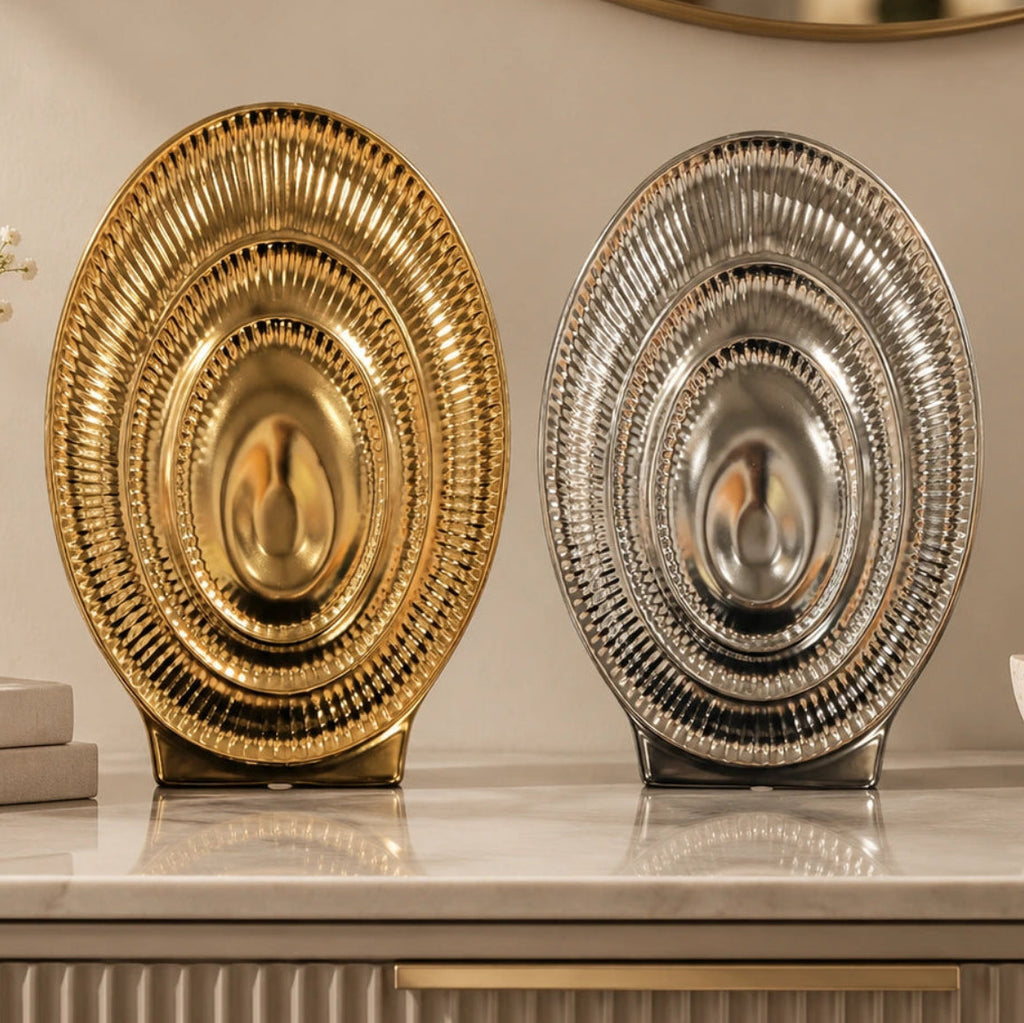 Two decorative oval platters with a ribbed, sunburst design, one in polished gold and one in polished silver, displayed on a marble countertop.