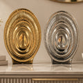 Two decorative oval platters with a ribbed, sunburst design, one in polished gold and one in polished silver, displayed on a marble countertop.