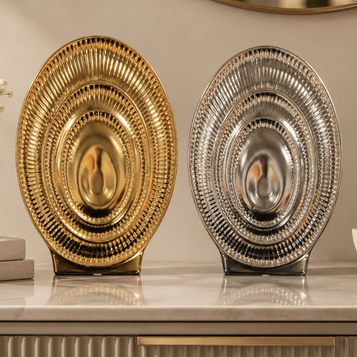 Two decorative oval platters with a ribbed, sunburst design, one in polished gold and one in polished silver, displayed on a marble countertop.