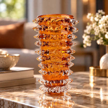 A tall amber glass vase with a fluted, tiered design and textured faceted details, displayed on a polished marble table in a warm, softly blurred living room setting.