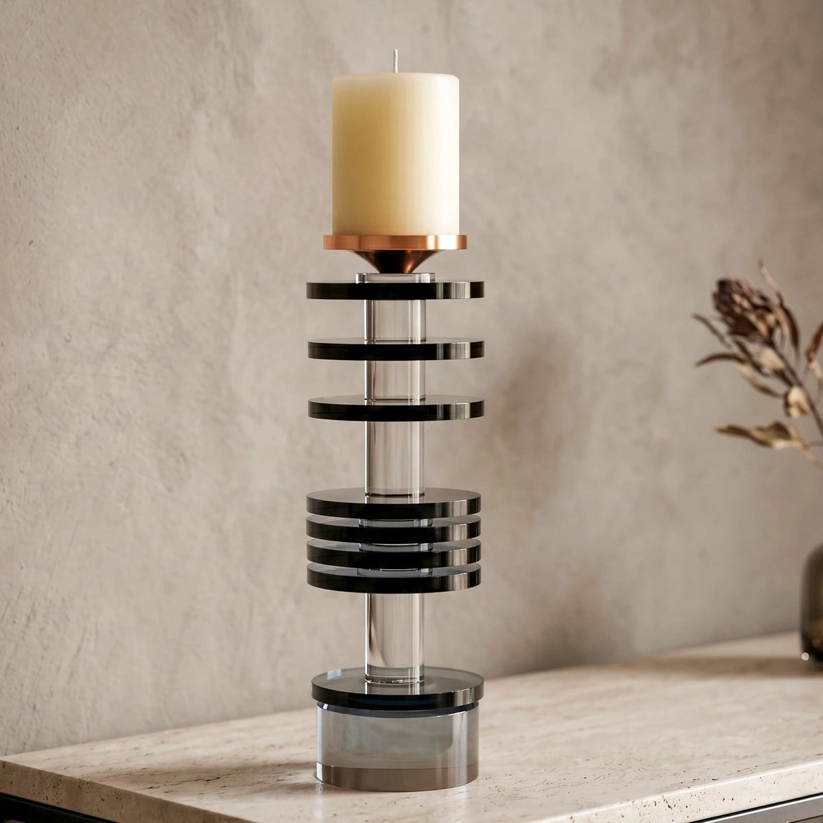 A modern geometric table candle holder with a cream pillar candle, featuring black and silver metallic rings and a copper-colored top band, displayed on a light stone surface against a textured beige wall.