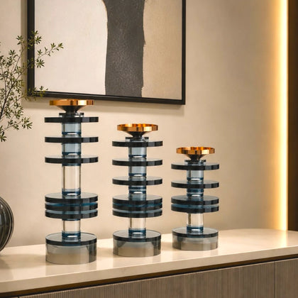 Three modern tiered glass candle holders with blue-gray translucent glass and gold-toned metal tops, arranged on a light console table in an interior setting.